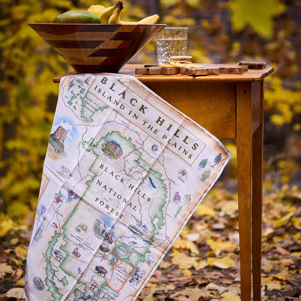 Map of Black Hills National Forest draped over a wooden table with a bowl of fruit and glass in an outdoor setting.