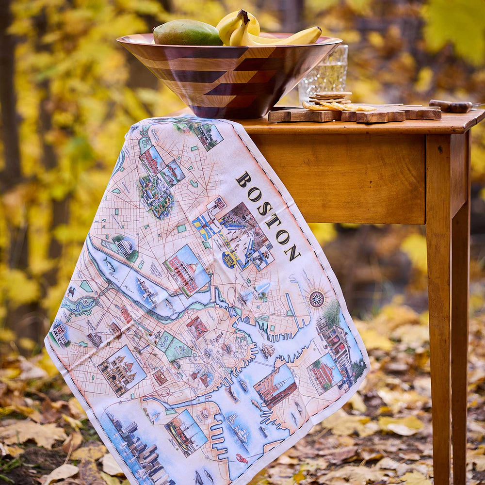Map of Boston towel draped over a wooden table with a bowl of fruit in an outdoor setting.