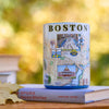 Candle with Boston design on top of a book against a blurred autumn background