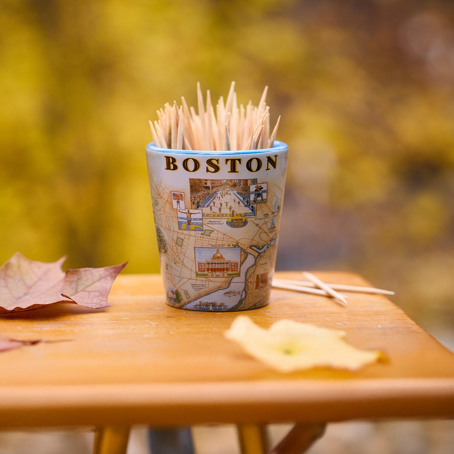 A Boston-themed toothpick cup filled with wooden toothpicks, sitting on a wooden table outdoors with autumn leaves and a blurred yellow foliage background.