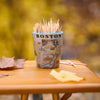 A Boston-themed toothpick cup filled with wooden toothpicks, sitting on a wooden table outdoors with autumn leaves and a blurred yellow foliage background.