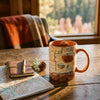 Map-themed mug on a wooden table with a notebook and pine cone, near a window with a forest view.