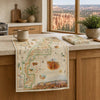 A vintage-style map table runner featuring Grand Canyon National Park is draped over a wooden kitchen counter, with a matching folded napkin and a ceramic mug nearby. A large window behind shows a scenic canyon view.