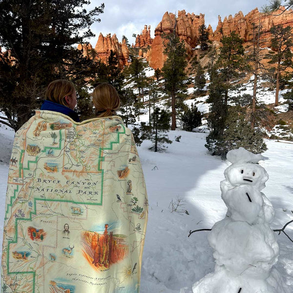 Two people wrapped in a Bryce Canyon fleece blanket next to a snowman. The backdrop is Bryce Canyon National Park red hoodoos.. 