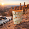 A cylindrical travel mug printed with an antique map of Bryce Canyon National Park sits on a rocky ledge outdoors at sunset, with folded sunglasses and a map nearby and hoodoos in the background.
