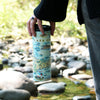 A person carrying the Cape Cod travel drinkware by the top while walking in a creek with large rocks.