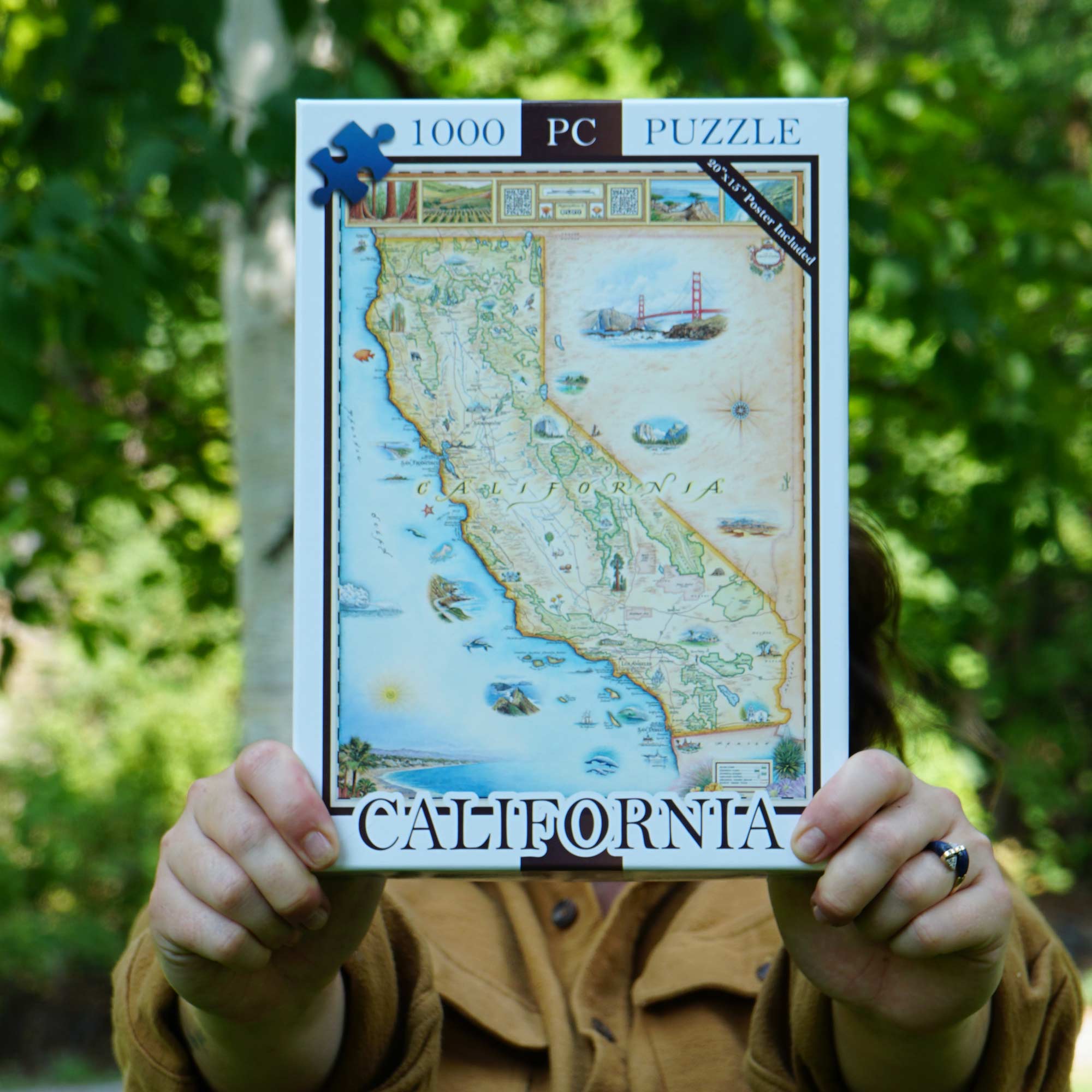 A person outdoors holds up a 1000-piece California jigsaw puzzle box, with their hands and torso visible against a blurred green forest background.