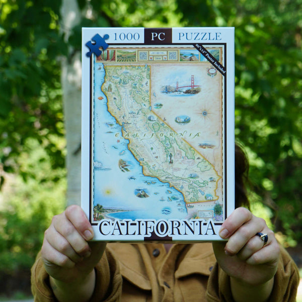 A person outdoors holds up a 1000-piece California jigsaw puzzle box, with their hands and torso visible against a blurred green forest background.