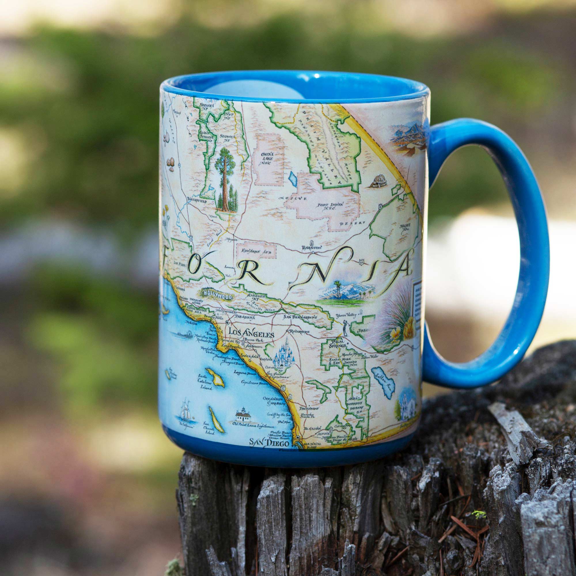 Blue Southern California coffee cup sitting on a tree stump. The cup featuring Los Angeles, pacific ocean, San Diego, Hollywood sign, and redwoods.