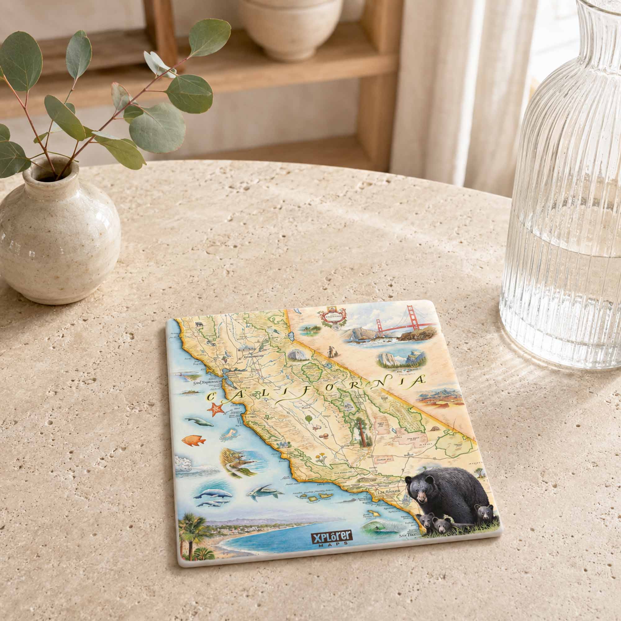 A square ceramic tile coaster featuring a vintage-style map of California with illustrated marine and terrestrial details, displayed on a beige stone tabletop next to a small vase and a glass bottle.