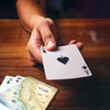 A close-up of a hand holding an Ace of Spades playing card over a wooden table, with two more cards partially visible in the foreground.