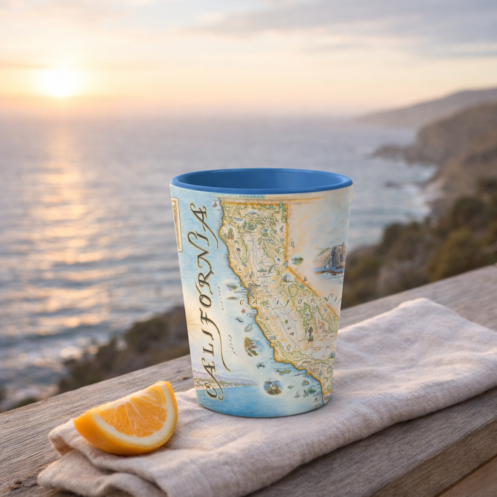 A ceramic tumbler with a blue interior and a vintage-style California map print sits on a wooden table outdoors, next to an orange slice and a folded beige cloth, with a blurred ocean sunset in the background.