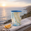 A ceramic tumbler with a blue interior and a vintage-style California map print sits on a wooden table outdoors, next to an orange slice and a folded beige cloth, with a blurred ocean sunset in the background.