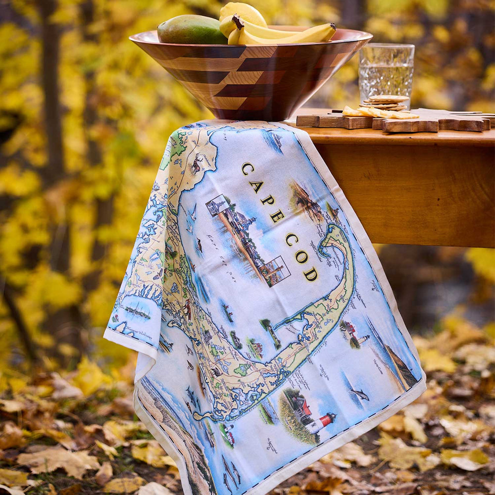 Cape Cod map towel draped over a wooden bowl with fruit and a glass on a table outdoors.