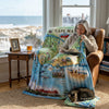 Woman reading a book on a Cape May-themed blanket with a cat next to her in a cozy room.