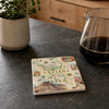 A square ceramic coaster featuring a vintage-style map of Capitol Reef National Park, styled on a dark gray kitchen countertop next to a potted plant and a glass pitcher of coffee.