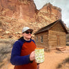 A person holding a Capitol Reef National Park Mug. In the background is a log cabin that is inside the park. 