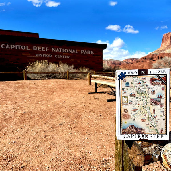 A jigsaw puzzle box labeled '1000 PC PUZZLE' and 'CAPITOL REEF' is shown in the foreground on a wooden post, with a Capitol Reef National Park sign and desert landscape in the background under a blue sky.