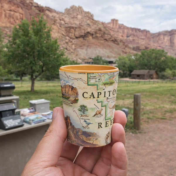 A hand holds a small illustrated cup featuring a vintage map design with the text 'CAPITOL REED' and scenic artwork, with a blurred outdoor desert canyon and grassy area in the background.