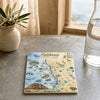 A square ceramic coaster featuring a colorful illustrated map of California's Central Coast State Parks, styled on a stone tabletop next to a potted plant and a glass carafe.