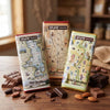 Three Xplorer Maps milk chocolate bars with vintage map packaging are arranged on a wooden table with scattered chocolate pieces, almonds, and cinnamon sticks. The background is a softly blurred rustic indoor setting.