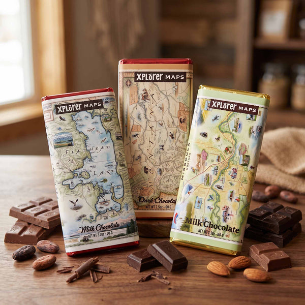 Three Xplorer Maps milk chocolate bars with vintage map packaging are arranged on a wooden table with scattered chocolate pieces, almonds, and cinnamon sticks. The background is a softly blurred rustic indoor setting.
