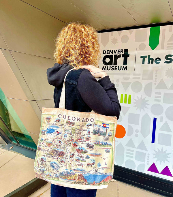 A woman standing in front of the Denver Art Museum carrying a canvas tote bag with a map of Colorado that showcases various cities including Denver, Fort Collins, Colorado Springs, Aspen, and Durango. It also features local flora and fauna, such as moose, Rocky Mountain elk, turtles, eagles, and Bighorn Sheep, as well as the indigenous Navajo and Hopi peoples. The map highlights popular activities like hiking, biking, rafting, and skiing.