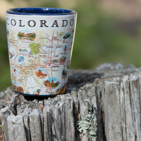Xplorer Maps shot glass of Colorado sitting on a tree stump. Colorado Map showcases various cities including Denver, Fort Collins, Colorado Springs, Aspen, and Durango. It also features local flora and fauna, such as moose, Rocky Mountain elk, turtles, eagles, and Bighorn Sheep, as well as the indigenous Navajo and Hopi peoples. The map highlights popular activities like hiking, biking, rafting, and skiing. 