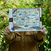 A person in a brown shirt holds up a 1000-piece jigsaw puzzle box featuring a colorful illustrated map of Colorado. The background is a blurred outdoor garden with green foliage and small red flowers.