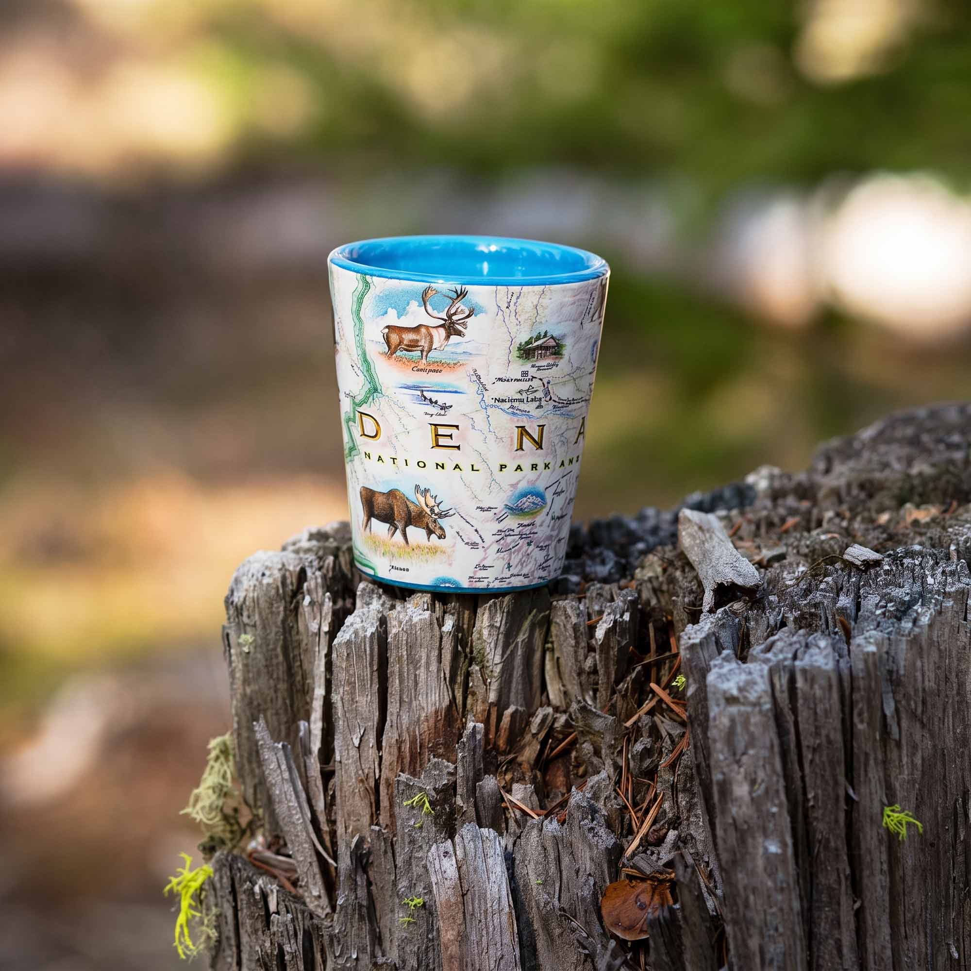 A ceramic shot glass featuring a map of Denali National Park with illustrations of moose and a blue interior rim, resting on a weathered tree stump in an outdoor forest setting.