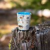 A ceramic shot glass featuring a map of Denali National Park with illustrations of moose and a blue interior rim, resting on a weathered tree stump in an outdoor forest setting.