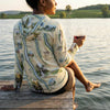 Person sitting on a dock by a lake, wearing a sun shirt hoodie with a Finger Lakes map design, holding a glass of wine.