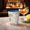 A ceramic shot glass featuring a colorful illustrated map of Finger Lakes, set on a marble countertop next to a cocktail in a coupe glass and fresh lemon slices, with a blurred bar background.