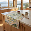 A rectangular kitchen table runner with a vintage-style illustrated map design draped over a light wood island in a bright kitchen. The map features pastel blue, green, and beige tones with small illustrations of landmarks, birds, and animals.