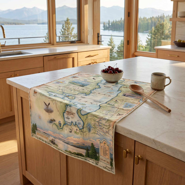 A rectangular kitchen table runner with a vintage-style illustrated map design draped over a light wood island in a bright kitchen. The map features pastel blue, green, and beige tones with small illustrations of landmarks, birds, and animals.