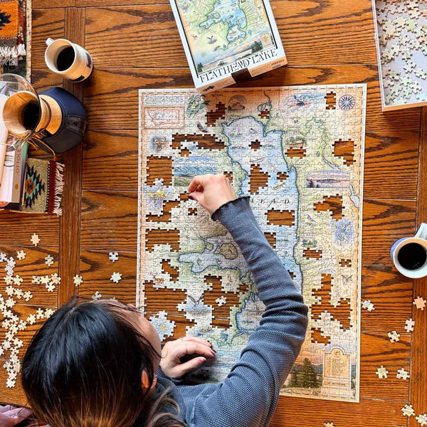 Person working on a puzzle of a map on a wooden table with coffee and books.