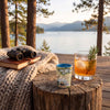 A styled outdoor lakeside scene on a wooden deck with a tree stump table. A small illustrated tumbler cup and a glass of amber cocktail with a rosemary garnish sit on the stump, alongside a knitted blanket, binoculars on a book, and a scenic lake and mountain background.