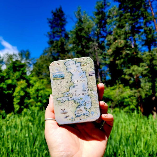 Flathead Lake map tin carrying case. The background is a forest with tall green grass. 