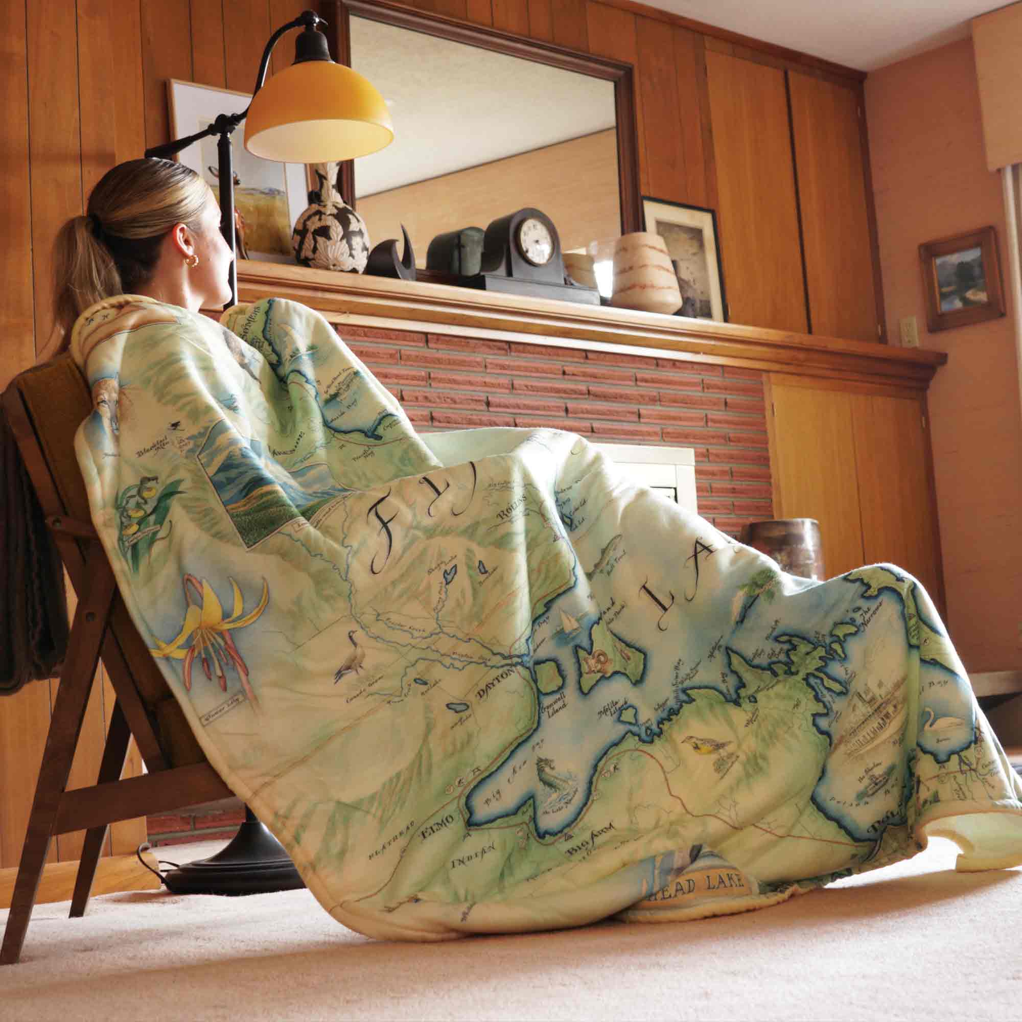 Woman in cabin sitting with Flathead Lake blanket covering her from head to toe. Hand-drawn map artwork on folded blanket, measuring 58