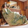 Woman in cabin sitting with Flathead Lake blanket covering her from head to toe. Hand-drawn map artwork on folded blanket, measuring 58"x50", showcasing scenic beauty of Flathead Lake in Montana