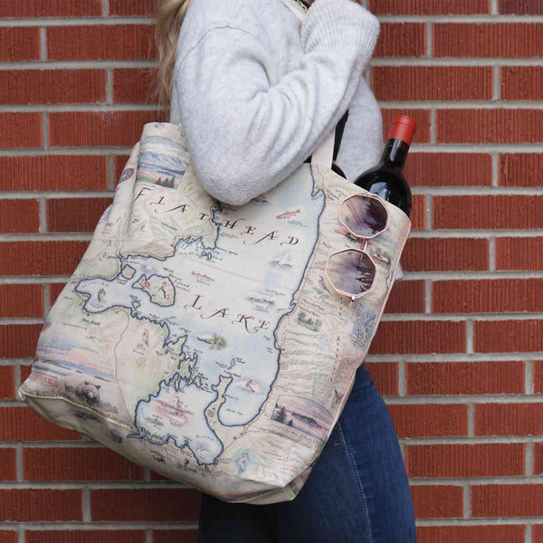 Woman with Flathead Lake Montana Map Canvas Tote Bag, carrying wine and sunglasses against brick wall backdrop. Map highlights activities like golfing, rafting, wildlife such as eagles, bears, deer, mountain lions, and flora like Glacier Lilies.