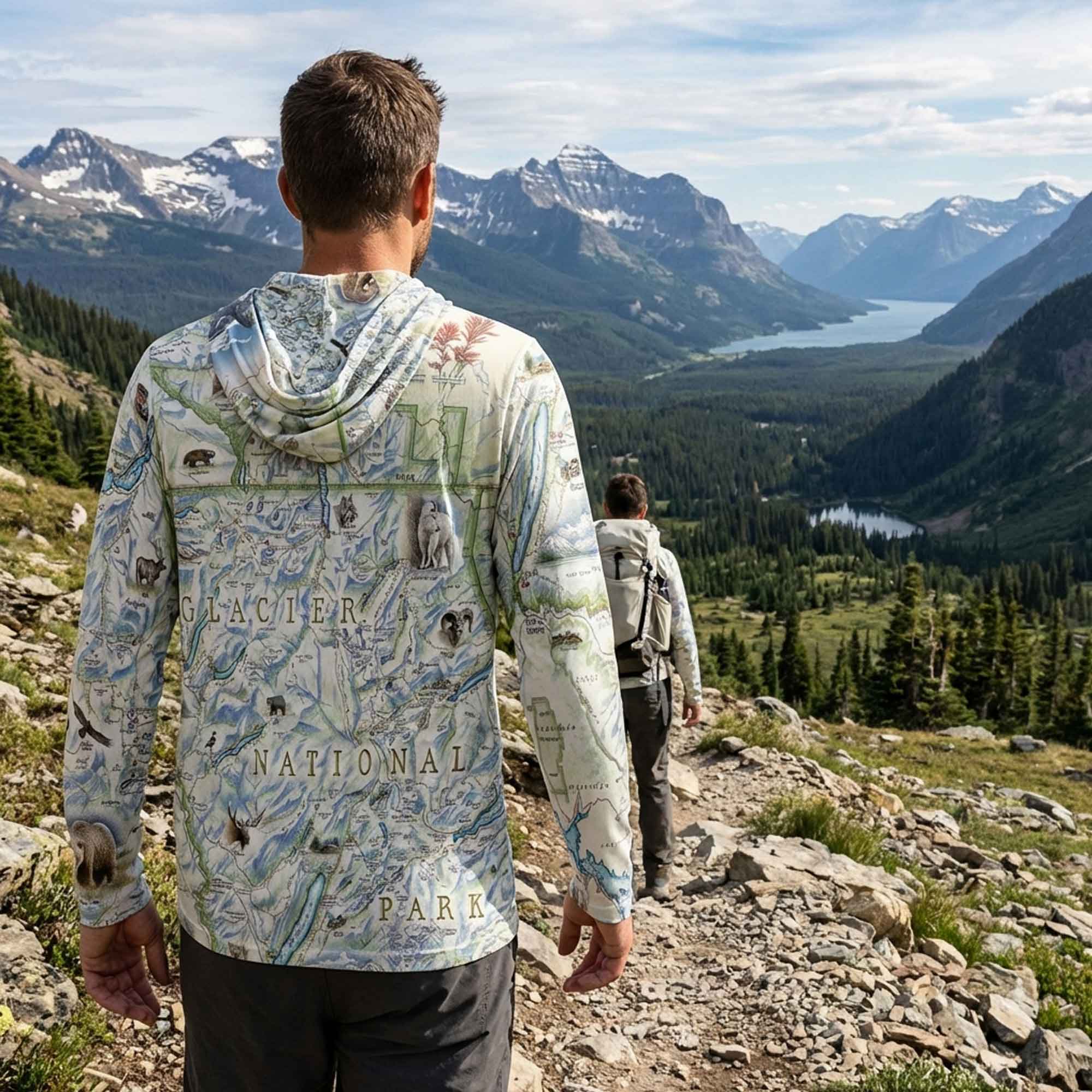 Person wearing a map-patterned sun shirt hoodie with Glacier National Park design, standing on a mountain trail with scenic view.