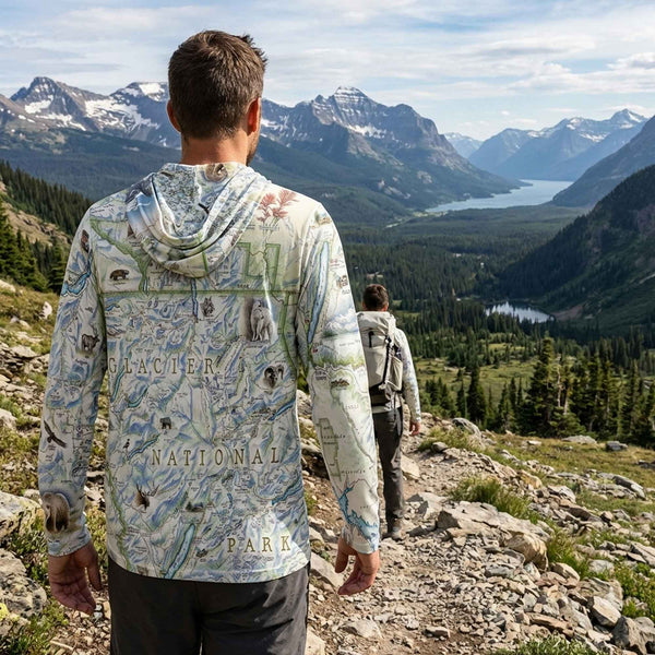 Person wearing a map-patterned sun shirt hoodie with Glacier National Park design, standing on a mountain trail with scenic view.