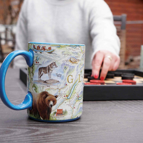 Background: Person playing checkers. Montana's Glacier National Park Ceramic Coffee Mug in blue, 16 oz, featuring mountains, goats, flowers, and more!