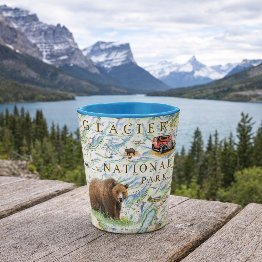 A ceramic cup featuring Glacier National Park artwork, including a brown bear, map details, and a red jeep, set on a wooden table with a scenic mountain lake background.