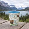 A ceramic cup featuring Glacier National Park artwork, including a brown bear, map details, and a red jeep, set on a wooden table with a scenic mountain lake background.