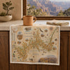 A rectangular table runner featuring an antique-style Grand Canyon National Park map print, styled on a kitchen counter with a mug and a folded matching napkin, with a window view of the canyon in the background.