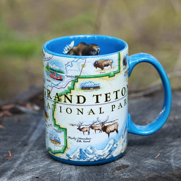 Grand Teton National Park Coffee Mug sitting on a log. The cup features moose, elk, bison, and mountains. Blue - 16 oz