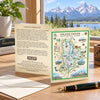 A Grand Teton National Park illustrated map card displayed on a wooden table, with its back side showing descriptive text. The card is styled with an envelope, a fountain pen, and a potted plant, with a scenic mountain lake background.
