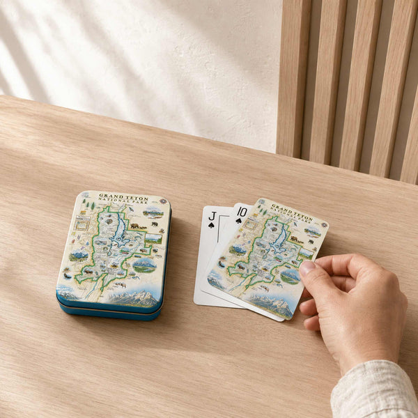 A hand is holding a Grandtown playing card deck next to its blue tin box on a light wood table. The cards and tin feature a detailed vintage-style map illustration of Grandtown with blue waterways and green landmasses.